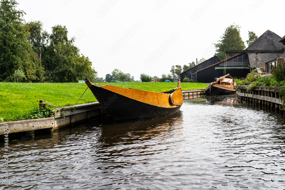 A wooden boat is moored in a canal by the shore, visible trees and buildings. A wooden boat is moored in a canal by the shore, visible trees and buildings.