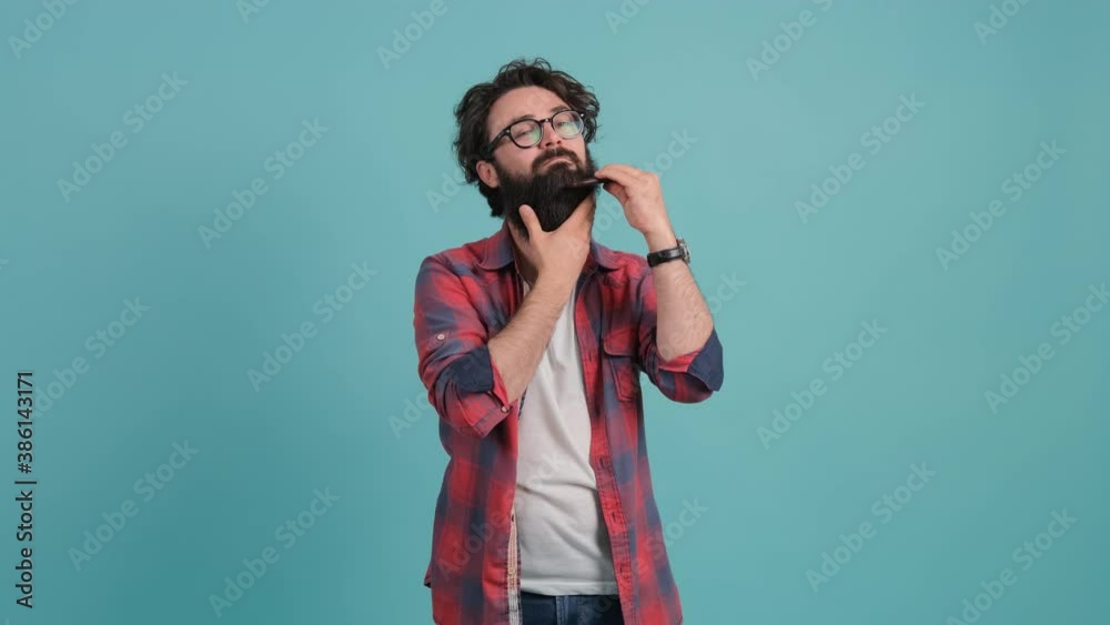 Young bearded man combing his beard looking at camera, isolated over turquoise background. Young bearded man combing his beard looking at camera, isolated over turquoise background.