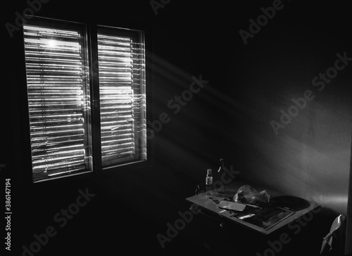 Dark small room in old apartment with wooden table and chairs near the window, sunlight coming through the blinds. Hard living background, depression and poverty concept. Black and white photography.