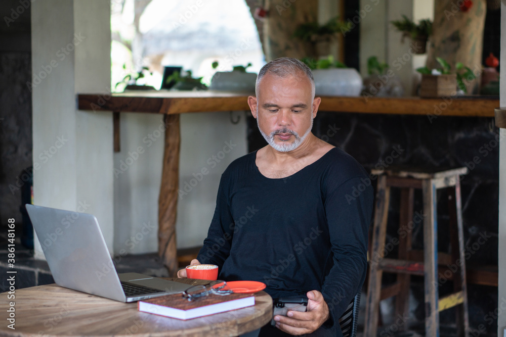 Mature man freelancer working with laptop and phone holding red cup of coffee Mature man freelancer working with laptop and phone holding red cup of coffee