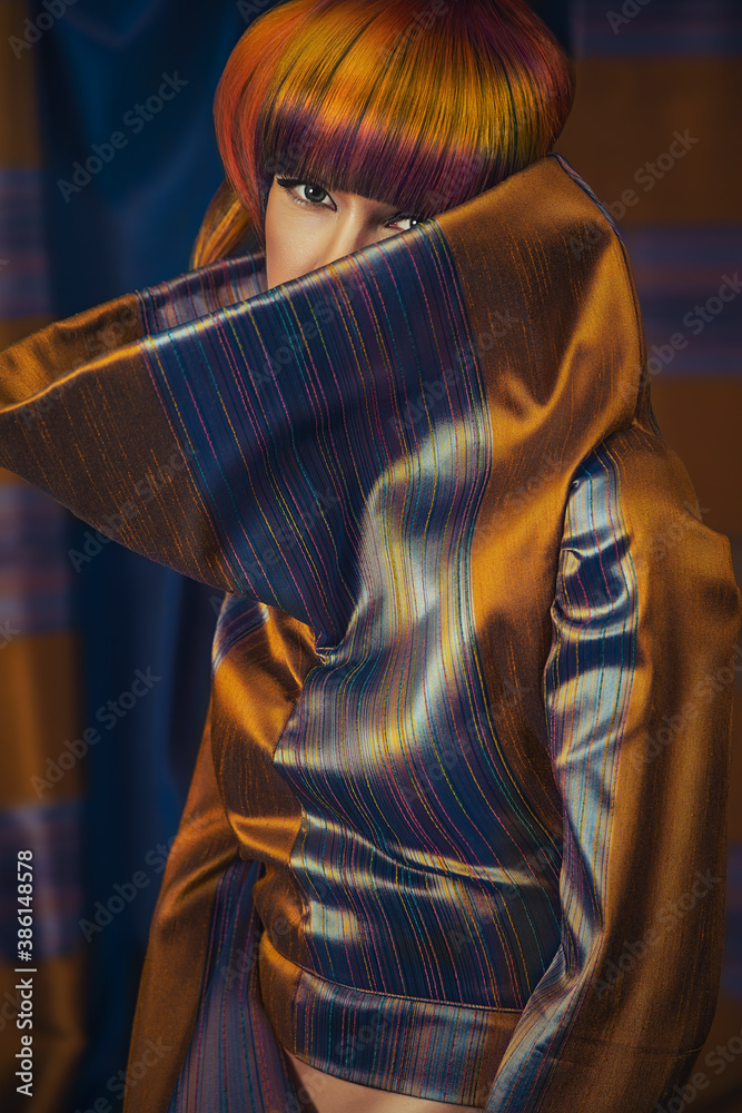 Fototapeta premium Portrait of a young woman wearing a copper and blue mock-neck outfit, posing with a colorful hairstyle while looking at the camera