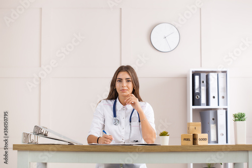 Woman pediatrician in her light office at the table in a white coat with a stenantoscope