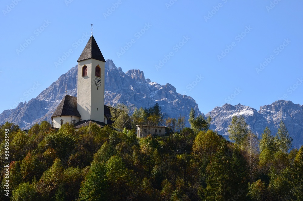 Dolomites mountains near the village of Innichen in South Tirol (Alto Adige), Italy, a traditional church on a forested hill in front, colorful autumn landscape, blue sky background, a sunny day