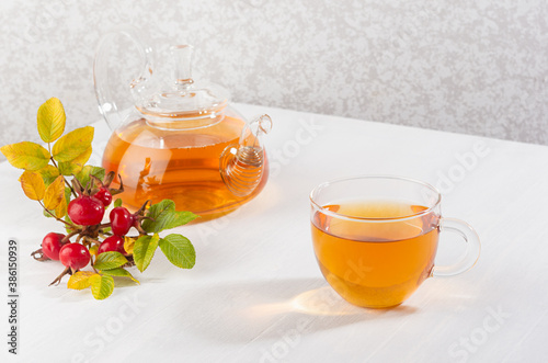 Cup of rose hip tea with teapot and autumn ripe dog rose branch on white wood table with copy space.