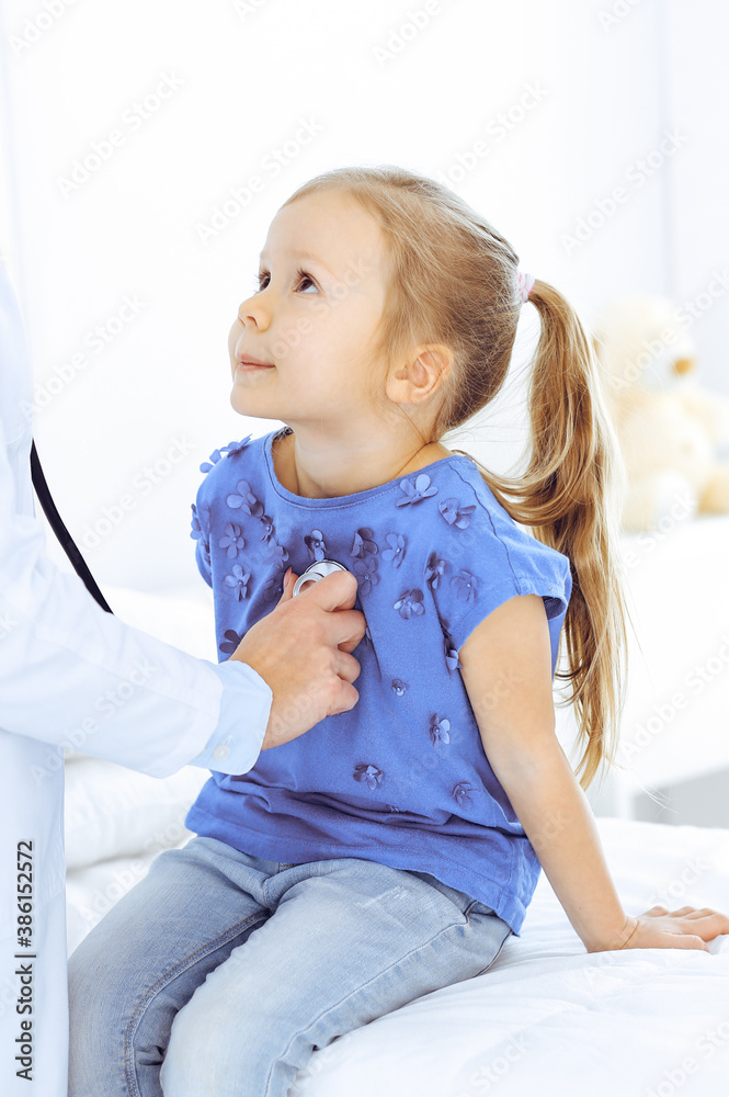 Fototapeta premium Doctor examining a little girl by stethoscope. Happy smiling child patient at usual medical inspection. Medicine and healthcare concepts