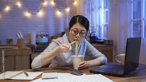 Fotografi busy asian chinese businesswoman eating unhealthy fast food sitting at dining table while looking at paper by laptop computer