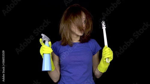 A young woman with tousled hair finished cleaning. Woman in safety glasses and gloves with tools for cleaning the toilet. Girl holds a toilet brush and water spray. On a black background