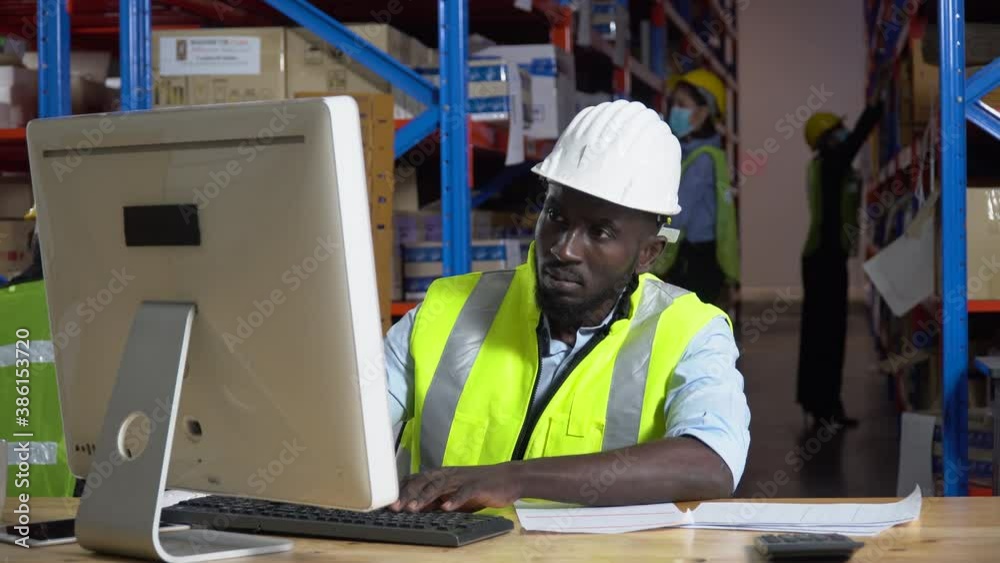 african warehouse manager sitting at desk using computer check inventory items online at distribution warehouse. black man worker in Hard Hat typing keyboard entering data document in workplace african warehouse manager sitting at desk using computer check inventory items online at distribution warehouse. black man worker in Hard Hat typing keyboard entering data document in workplace