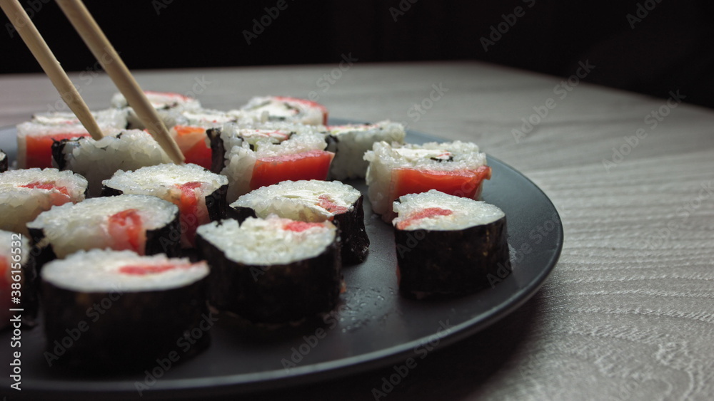 Smooth hit on a plate with Japanese food. Female hand with chopsticks takes sushi from a plate.Sushi rolls japanese food rotated over black background. California Sushi roll with tuna, vegetables Smooth hit on a plate with Japanese food. Female hand with chopsticks takes sushi from a plate.Sushi rolls japanese food rotated over black background. California Sushi roll with tuna, vegetables