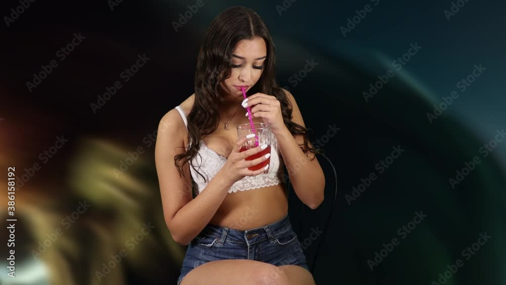 Eine junge schöne Frau in einem weißen BH und dunklem  kurzen Jeansrock trink verführerisch aus einem Glas durch einen Strohhalm 