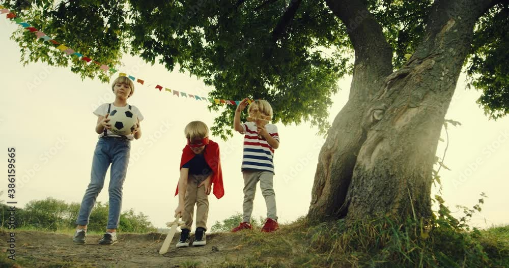 Portrait shot of the three Caucasian teen little boys playing at the big tree in summer- one in the red mask and mantle of superhero with a sword like a knight, one with a football ball and one in the Portrait shot of the three Caucasian teen little boys playing at the big tree in summer- one in the red mask and mantle of superhero with a sword like a knight, one with a football ball and one in the