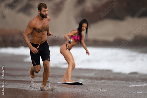 A young man pulls a young girl on a rope who is learning a skimboard