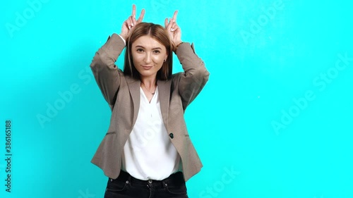 Adult woman, arrogant, stubborn, in business style jacket, imitating horns, holding fingers above her head, dangerous behavior of boss, domineering. Studio image indoors, isolated on blue background