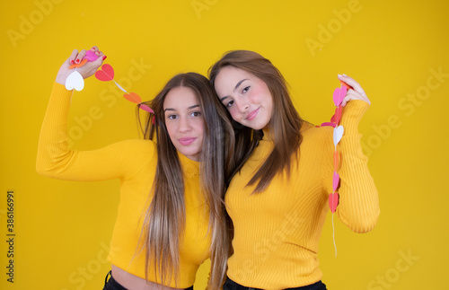 Portrait of two cheerful young girls standing together and looking at camera isolated over yellow background