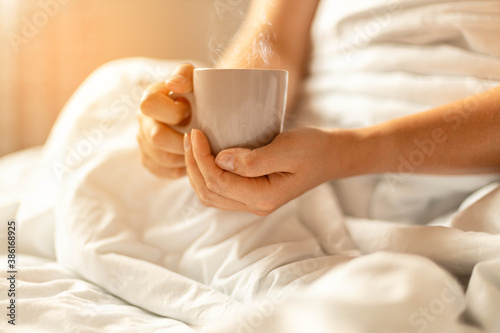 Woman hand holding cup of coffee in bed on white blanket.