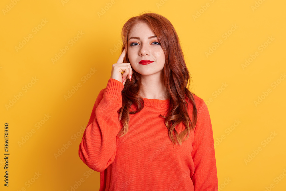 Young adult girl posing with pensive look, holding finger on temple. Attractive woman in orange sweater having new great idea, looks at camera, posing isolated over yellow wall. Young adult girl posing with pensive look, holding finger on temple. Attractive woman in orange sweater having new great idea, looks at camera, posing isolated over yellow wall.