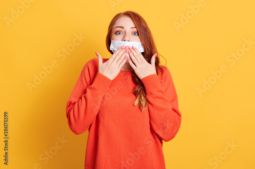Young woman covering mouth with hand, looks shocked with shame for mistake, expression of fear, looking directly at camera with big eyes, standing against yellow wall.