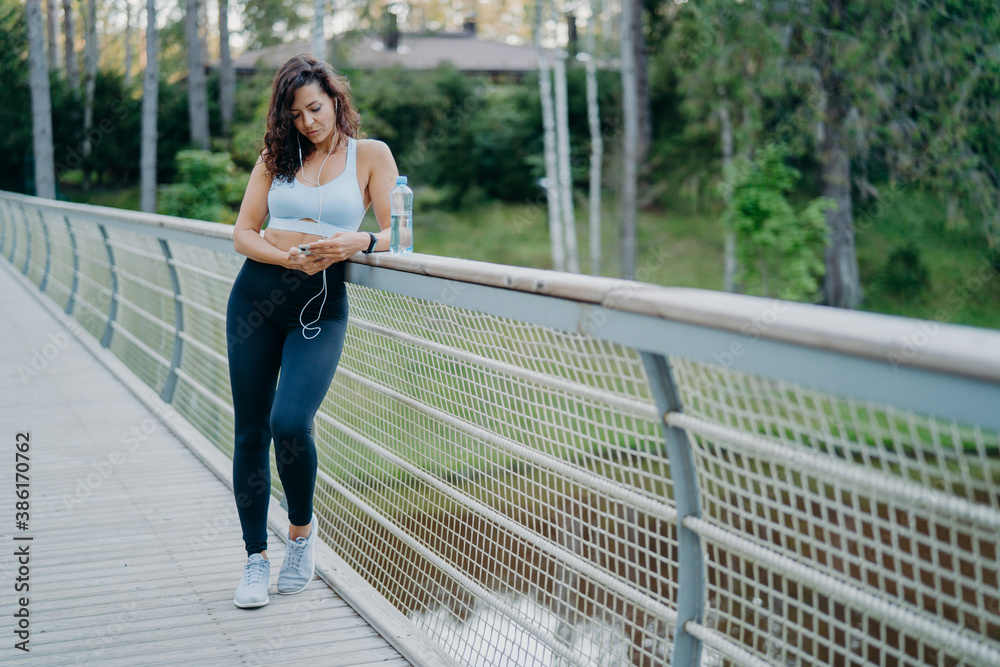 Sporty slim European woman in active wear checks notification on mobile phone, listens music from playlist uses earphones, poses on bridge against nature background. Sport and lifestyle concept