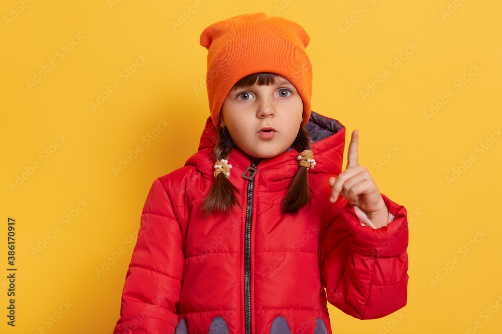 Caucasian preschooler girl shows index finger up isolated over yellow background, has great idea, has thoughtful look, wearing red jacket and orange cap. Caucasian preschooler girl shows index finger up isolated over yellow background, has great idea, has thoughtful look, wearing red jacket and orange cap.