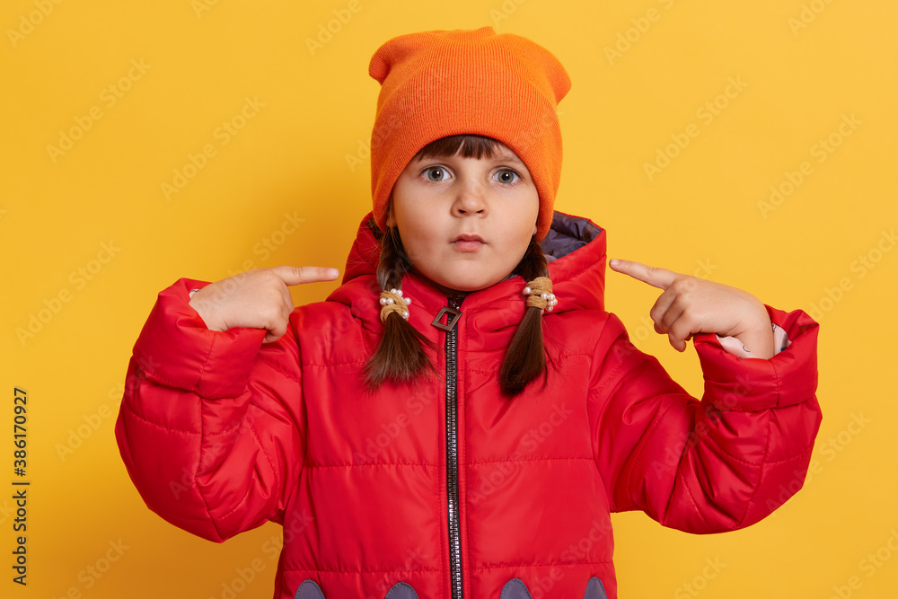 Small girl looking at camera with big eyes, pointing at her cheeks with her index fingers, making funny grimace with her lips isolated over yellow background, kid wearing red jacket and orange cap. Small girl looking at camera with big eyes, pointing at her cheeks with her index fingers, making funny grimace with her lips isolated over yellow background, kid wearing red jacket and orange cap.