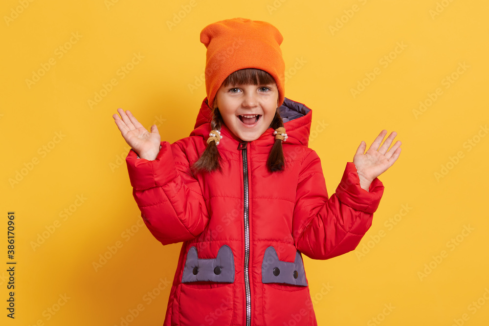 Cute smiling little child raising her hands up isolated on yellow background, looks at camera with opened mouth, expressing excitement, kid with pigtails dresses casual jacket and cap. Cute smiling little child raising her hands up isolated on yellow background, looks at camera with opened mouth, expressing excitement, kid with pigtails dresses casual jacket and cap.