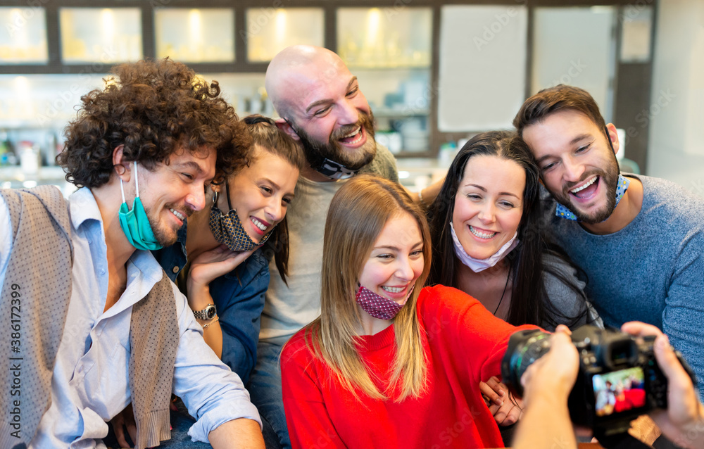 Obraz premium smiling group of caucasian people wearing protective face mask posing for a picture together in front of a photographer reflex camera. mates taking a pic together. concept about new normal lifestyle