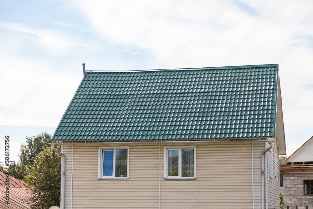 The roof of an old wooden house covered with sheets of green metal tiles on a background of green coniferous trees on a summer day. Business selling building materials or helping low-income families.