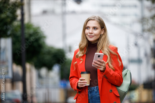 Cheerful young woman wearing coat walking with coffee cup and smartphone