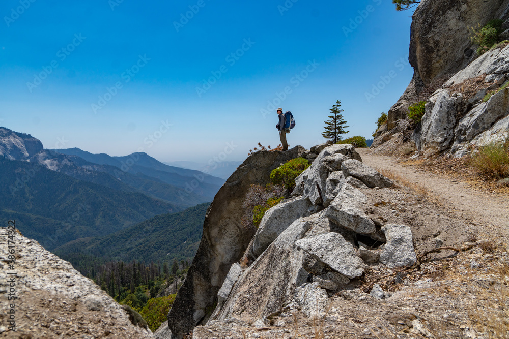 A Hiker standing on the edge of a cliff by a trail, high above a valley below and distance mountains, Sequoia NP, California