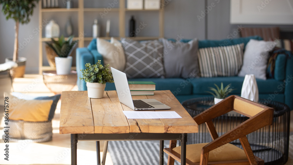 Sunlight illuminates living room workplace home office interior for comfort productive work, brown and blue colours. On wooden table laptop comfy couch with cushions on background, workday end concept