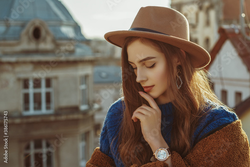 Outdoor fashion portrait of young elegant fashionable brunette woman, model wearing stylish hat, wrist watch, brown faux fur coat, posing at sunset, in European city. Copy empty space for text
