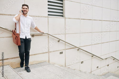 Positive young man standing on steps and talking on phone with friend or coworker
