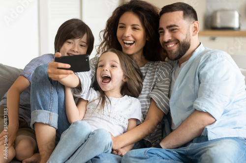 Overjoyed parents and two adorable kids posing for selfie together, cute smiling girl holding smartphone, photographing, happy mother and father with children having fun with gadget at home