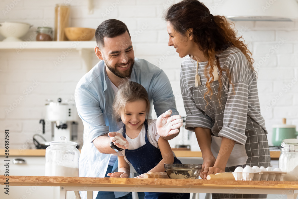 Happy parents with adorable little daughter having fun with dough, baking pastry or pie together, laughing father clapping hands with flour, overjoyed family enjoying leisure time in kitchen Happy parents with adorable little daughter having fun with dough, baking pastry or pie together, laughing father clapping hands with flour, overjoyed family enjoying leisure time in kitchen