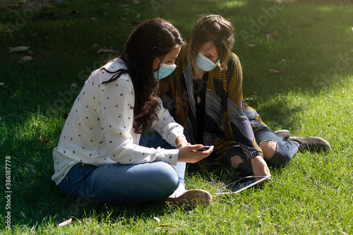 Couple of friends with medical mask, sitting in the park using their smartphone and tablet during the coronavirus pandemic