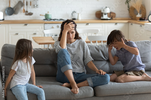 Tired exhausted mother having problem with naughty noisy kids, sitting on couch, touching forehead, suffering from headache, difficult child, children tantrum and manipulation concept