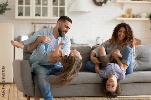 Happy young parents playing with two little kids at home, sitting on cozy couch, overjoyed mother and father holding adorable son and daughter upside down, tickling, family enjoying leisure time