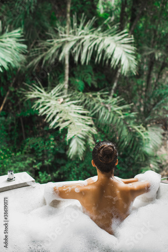 Woman relaxing in bath tub full of foam outdoors with jungle view. View from behind. Unrecognizable person. Beauty spa treatment, leisure time.