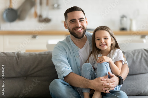 Portrait smiling father and little daughter hugging, looking at camera, sitting on cozy couch at home, happy young dad and cute adorable girl posing for family photo, enjoying leisure time together