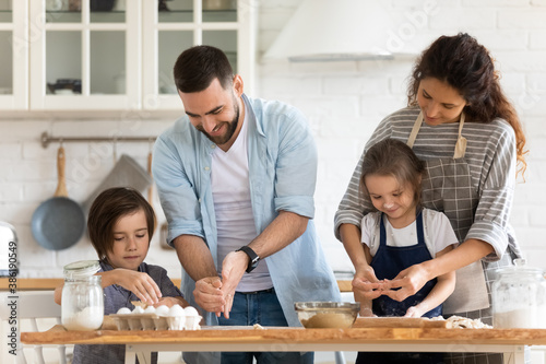 Happy mother and father with two kids baking together, standing at wooden countertop in modern kitchen, young parents with little daughter and son cooking pastry or pie, enjoying leisure time