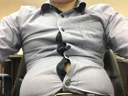 Man with big and fat belly wearing too small and tight shirt sitting on the chair. Need exercise for body fit. Healthcare and diet concept