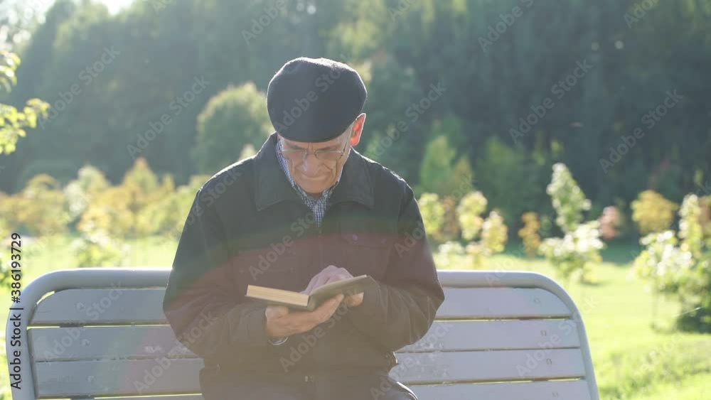 old man wearing glasses and cap reading book sitting on wooden bench in city park outdoor
