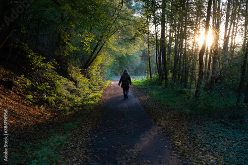 Woman walking on a road through a forest, the low sun shining through the trees.