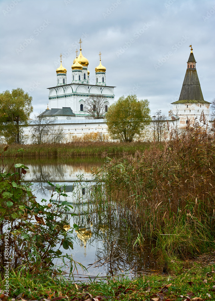 An ancient Russian monastery behind strong fortress walls with a pond in the foreground and the reflection of sparkling golden domes in the water. Landscape