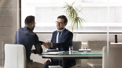 Smiling 30s businessman in glasses and suit shaking hands with happy arabic ethnic young male colleague, establishing partnership or making agreement after project negotiations in modern office.