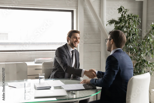 Happy young manager shaking hands with leader, feeling thankful for new job offer. Satisfied with good working results ceo executive manager praising male employee at meeting, sitting at table.