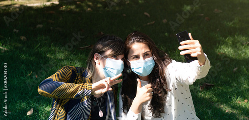 Couple of friends smiling behind the face mask, take a selfie with the smartphone while making the symbol of victory with their hands. Happy friends have a good time in the park during the new normal.