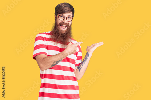 Handsome young red head man with long beard wearing casual clothes amazed and smiling to the camera while presenting with hand and pointing with finger.