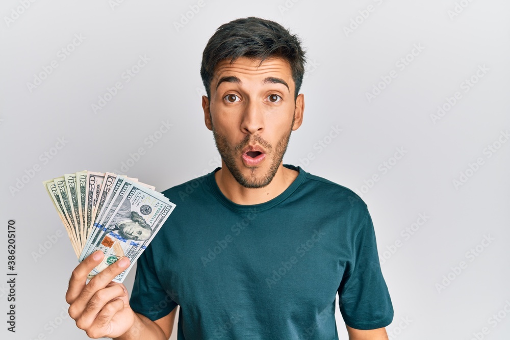 Young handsome man holding dollars scared and amazed with open mouth for surprise, disbelief face Young handsome man holding dollars scared and amazed with open mouth for surprise, disbelief face