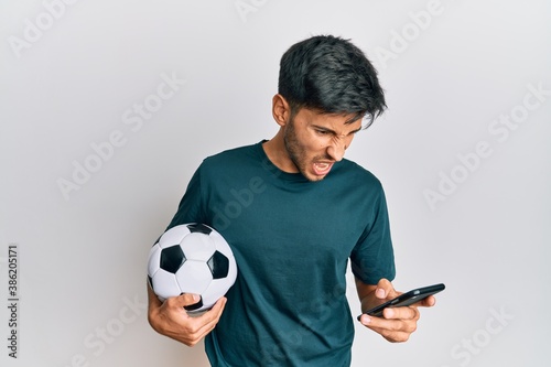 Young handsome man holding football ball looking at smartphone bet app angry and mad screaming frustrated and furious, shouting with anger. rage and aggressive concept. photo